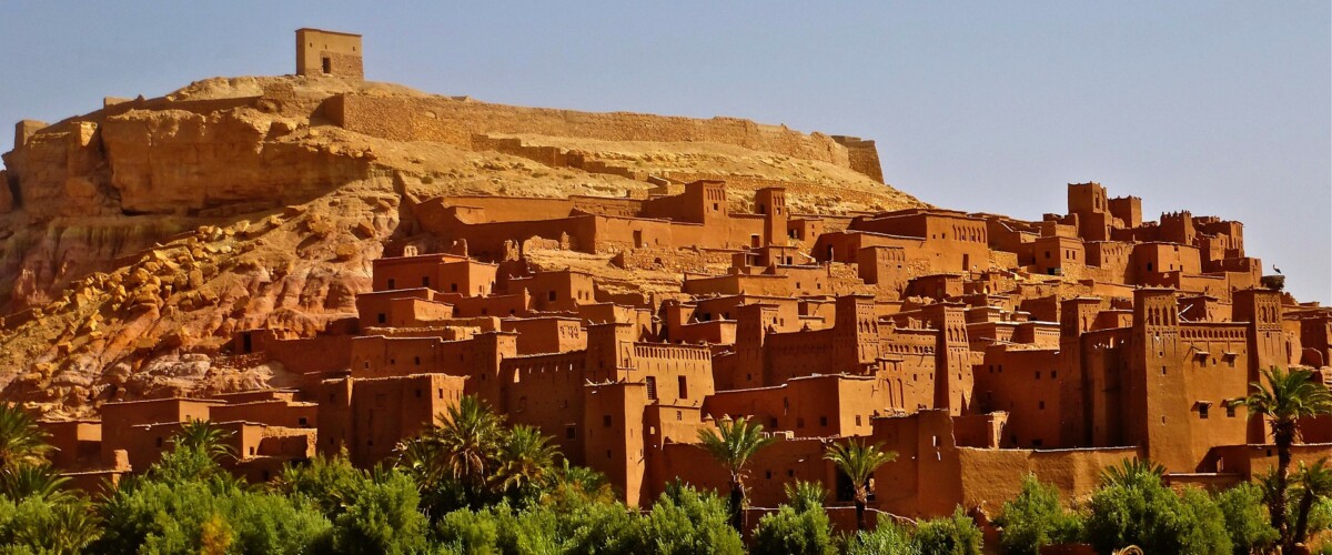 Kasbah di Ait Benhaddou con edifici in terra ocra arroccati su collina, fortezza in cima e vegetazione verde in primo piano nel deserto marocchino