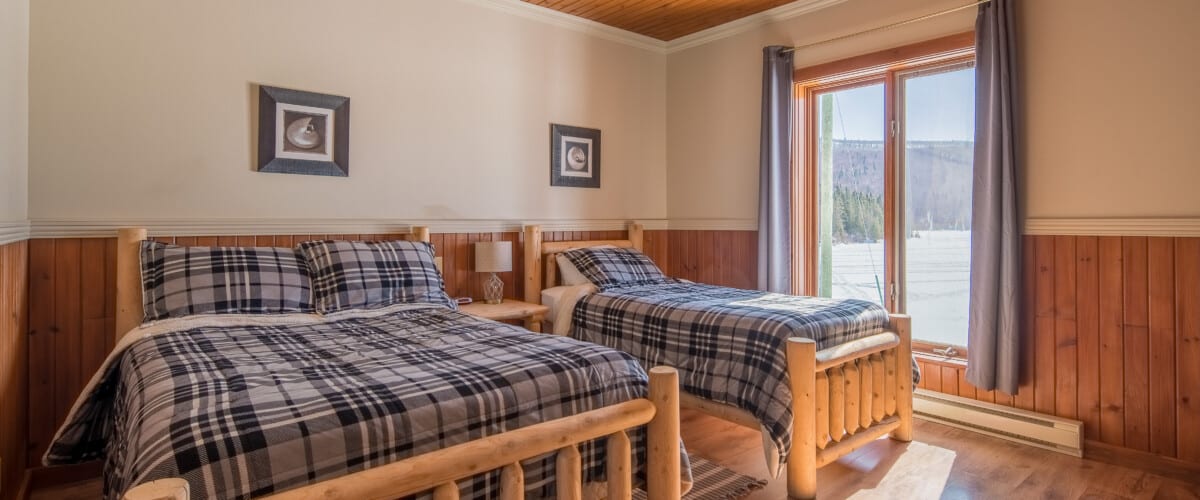 Camera da letto con due letti in legno, coperte scozzesi blu, finestre con vista sulle montagne innevate e pavimento in legno.