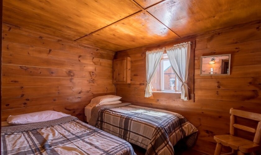 Camera rustica in legno con due letti singoli, finestra con tende bianche e arredi in legno naturale