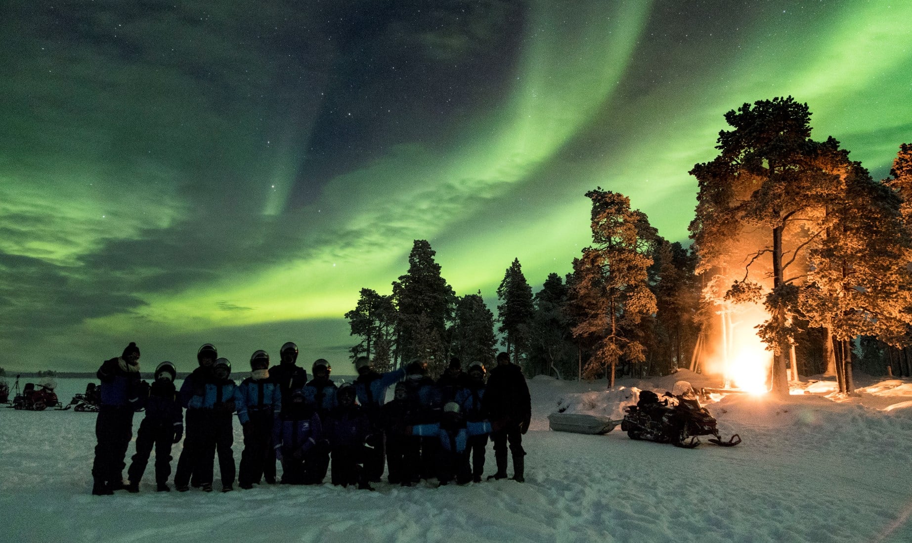 Gruppo di persone osserva l'aurora boreale verde sopra la foresta innevata della Lapponia con fuoco acceso a destra