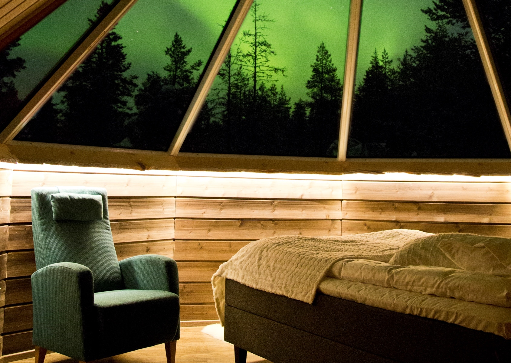 Interno di una kota in legno con poltrona verde e divano, vista notturna dell'aurora boreale verde attraverso il soffitto trasparente